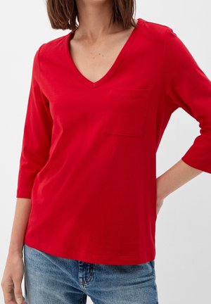 Vrouw draagt een rood topje met V-hals, driekwart mouwen en een borstzak, gecombineerd met een blauwe spijkerbroek.