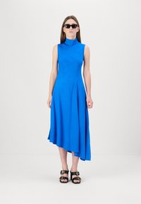 Asymmetrisches blaues Kleid mit hohem Kragen, ärmellosem Design und glatter Textur, kombiniert mit schwarzen Sandalen mit Schnallen-Details.