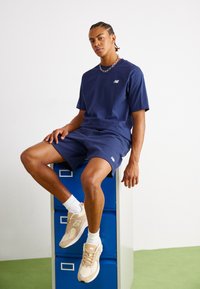 T-shirt in cotone blu navy e pantaloni corti, presenta il logo New Balance bianco. Sneakers atletiche beige e calze bianche. Modello seduto su un armadietto blu.