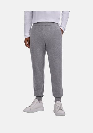 Persona con pantalones jogger de punto gris con puños acanalados, camiseta blanca de manga larga y zapatillas blancas, de pie sobre un fondo liso.