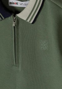 Polo vert texturé avec demi-fermeture éclair, col rayé, et petit logo brodé sur la poitrine indiquant "CMXT 97".