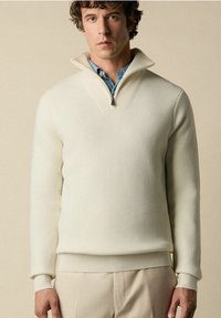 Bílý pulovr z ribovaného pleteného materiálu s vysokým límcem na zip, vrstvený přes modrou košili. Přiléhavý design s hladkou, měkkou strukturou.