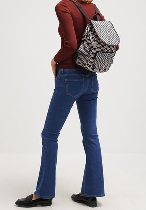 Schwarz-weiß gemusterter Rucksack mit Klappe, getragen von einer Person in einem burgunderroten Oberteil und blauen, ausgestellten Jeans. Glatte Textur und mittlere Größe.