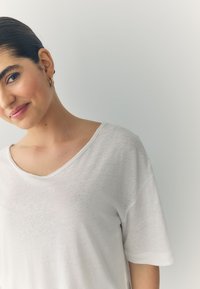 White, short-sleeved t-shirt with a V-neck design. Lightweight, slightly textured fabric. Model with slicked-back hair, posing against a light background.