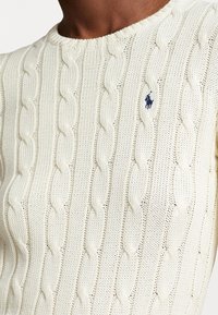 Krämfärgad kabelstickad crewneck tröja med liten marinblå broderad polo-spelare logotyp på bröstet mot mörk hudton.