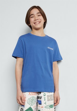 Blue cotton t-shirt with a crew neck and short sleeves, featuring a small white logo on the chest. Paired with patterned shorts.