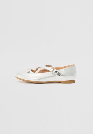 Chaussure plate Mary Jane argentée brillante avec une bride à boucle et un petit nœud décoratif sur une semelle marron clair, sur un fond gris uni.