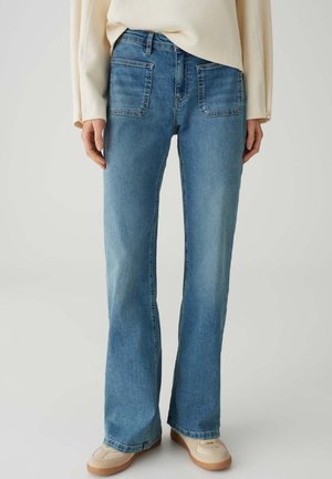 Femme portant un jean droit bleu clair avec des poches plaquées à l'avant, associé à des baskets beiges et un haut crème à manches longues.