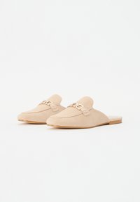 Muli bej din suede, slip-on, cu talpă plată, având o cataramă metalică decorativă și o textură netedă. Designul este minimalist și rotunjit.