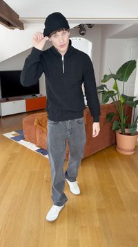 Black ribbed pullover with a zip neck, gray loose-fit jeans, and white sneakers. Indoor setting with a leafy plant and a brown couch.