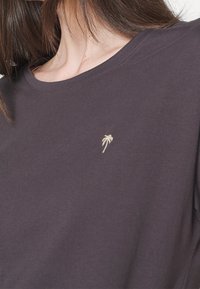 T-shirt en coton violet foncé avec un col rond. Présente une petite broderie de palmier beige sur la poitrine. Manches courtes, texture douce.