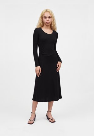 Une femme porte une robe midi noire à manches longues et à encolure arrondie, ainsi que des chaussures noires à talons et à brides, sur un fond blanc uni.