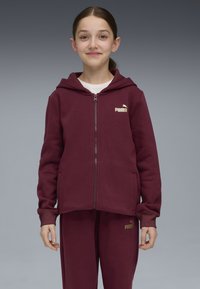 Felpa bordeaux con zip frontale, dettagli con tasche e un logo Puma dorato sul lato sinistro del petto, abbinata a pantaloni della tuta coordinati.