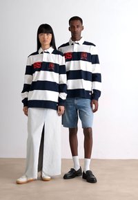 Marineblå og hvite stripete polo-skjorter med røde aksenter, kombinert med et langt denim-skjørt og shorts. Modellen står foran en nøytral bakgrunn.