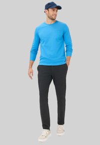 Blauw langemouwen shirt met een borstzak, gecombineerd met donkergrijze broeken en beige sneakers. De outfit heeft een casual, comfortabele uitstraling.