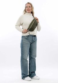 Borsa a tracolla in tessuto verde oliva con cerniera, indossata da una persona con un maglione bianco di tessuto strutturato e jeans blu, in piedi contro uno sfondo bianco.