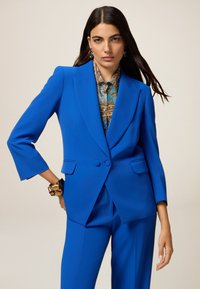 Blue suit with a fitted blazer, featuring a notched collar and two buttons. Trousers have a tailored fit. Printed shirt underneath adds contrast.