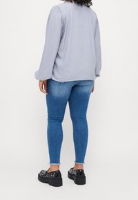 Blouse à manches longues gris clair avec des épaules froncées, associée à un jean skinny bleu. Des chaussures à plateforme noires avec des accents cloutés complètent le look.