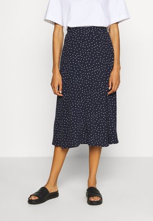 Falda midi azul marino con lunares blancos, confeccionada en un tejido suave, con forma acampanada y diseño de cintura alta. Combinada con sandalias negras tipo slide.