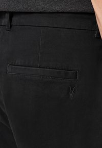 Nahaufnahme von schwarzen Hosen mit einer hinteren Paspeltasche und einem dezenten gestickten Ziegenkopf-Logo in der Nähe der Tasche.
