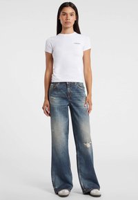 Witte t-shirt met korte mouwen en logo op de borst, gecombineerd met lichtblauwe, wijde distressed jeans en grijze sneakers.