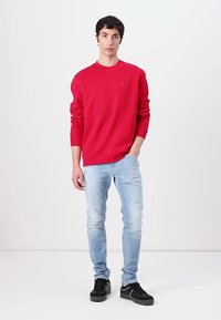 Rød sweatshirt med lange ærmer, rund halsudskæring og logo på brystet, parret med lyseblå jeans og sorte sneakers.