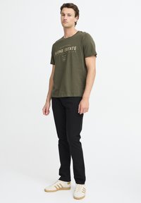 T-shirt en coton vert olive avec un imprimé doré, associé à un pantalon noir et des baskets blanches avec des rayures dorées. Design simple.