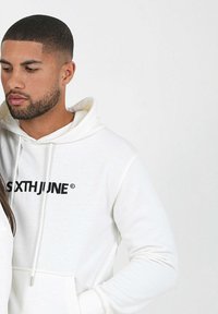 Sudadera blanca con bolsillo canguro frontal y logo negro "SIXTHJUNE". Tejido suave, capucha con cordón, textura suave, ajuste casual.