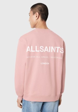 Man draagt een roze sweatshirt met "ALLSAINTS ACCESS ALL AREAS / BACKSTAGE LONDON" in het wit gedrukt op de achterkant, gecombineerd met lichtblauwe spijkerbroek.