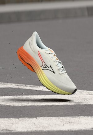 Chaussure de sport blanche avec semelle orange et jaune surélevée au-dessus de l'asphalte avec des marquages blancs au sol et de petits débris volant autour.