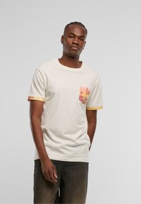 Camiseta de color crema con textura salpicada. Presenta un bolsillo con un patrón en tonos naranjas y rosas, y un borde amarillo en contraste en las mangas.