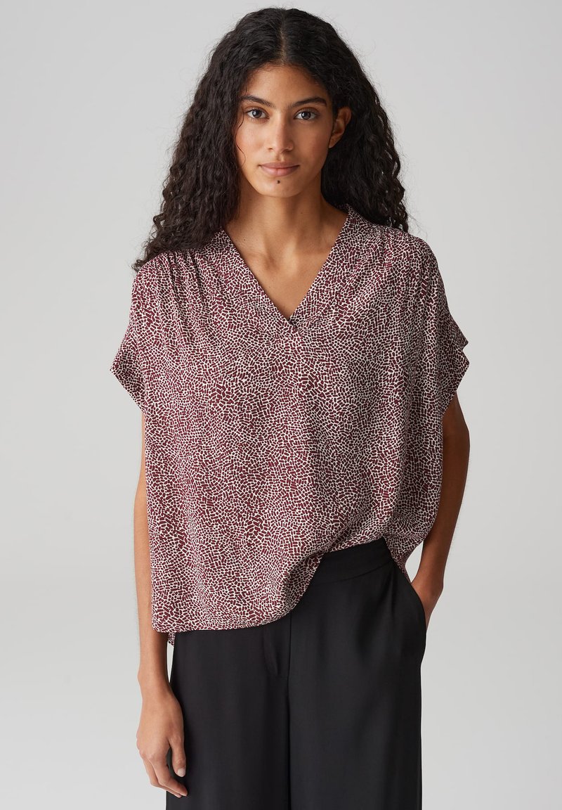 Blouse à manches courtes, décolletée en V profonde, en bordeaux avec un motif de points blancs texturé. Coupe loose qui se drape sur un pantalon noir, tissu doux.