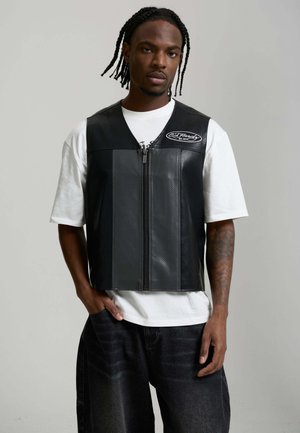 Mann mit geflochtenem Haar, der eine schwarze Lederweste mit Reißverschluss über einem weißen T-Shirt und schwarzen Jeans trägt, vor einem schlichten grauen Hintergrund.