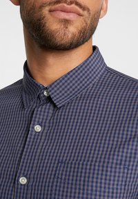 Camicia a quadri blu navy e grigia con colletto a punta, dotata di tasca frontale e bottoni in plastica rotondi. Materiale in cotone.