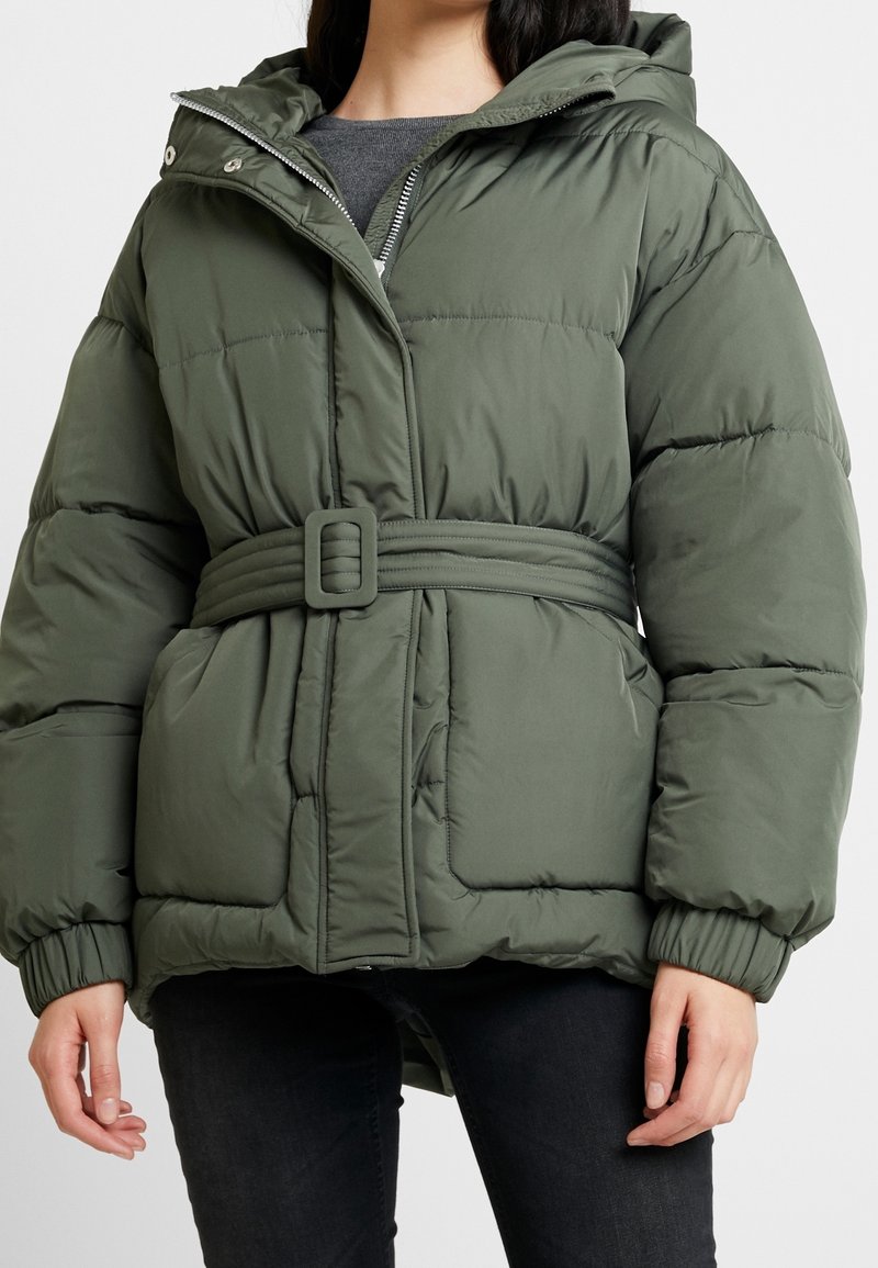 Kobieta w oliwkowo-zielonej, pikowanej kurtce z kapturem, paskiem i elastycznymi mankietami, ubrana w ciemne spodnie i szary top, stojąca na białym tle.