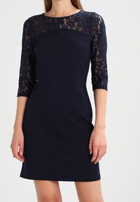 Robe bleu marine avec manches en dentelle trois-quarts et silhouette ajustée. Tissu lisse avec détails en dentelle aux épaules et aux bras.