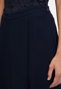 Granatowe, plisowane spodnie z gładkiego materiału, z paskiem w talii z dyskretnym zamkiem. Widoczna koronkowa bluzka o teksturowanej powierzchni.