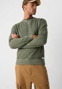 Groene geribbelde sweatshirt met ronde hals, met lange mouwen en een gestructureerde afwerking. Gecombineerd met kaki cargobroek, met subtiele branding.