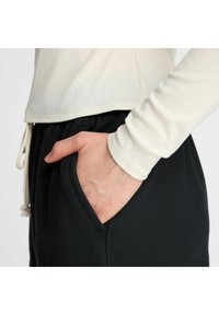 Beige geribde lange mouw top gecombineerd met zwarte sweatpants. De broek heeft zijzakken en een koord in de taille. Glad textuur.
