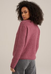 Puhasta, prevelik pulover v roza roza tkanini z teksturiranim pletenim vzorcem. Ima spuščena ramena in rebraste manšete, v kombinaciji z sivimi hlačami.