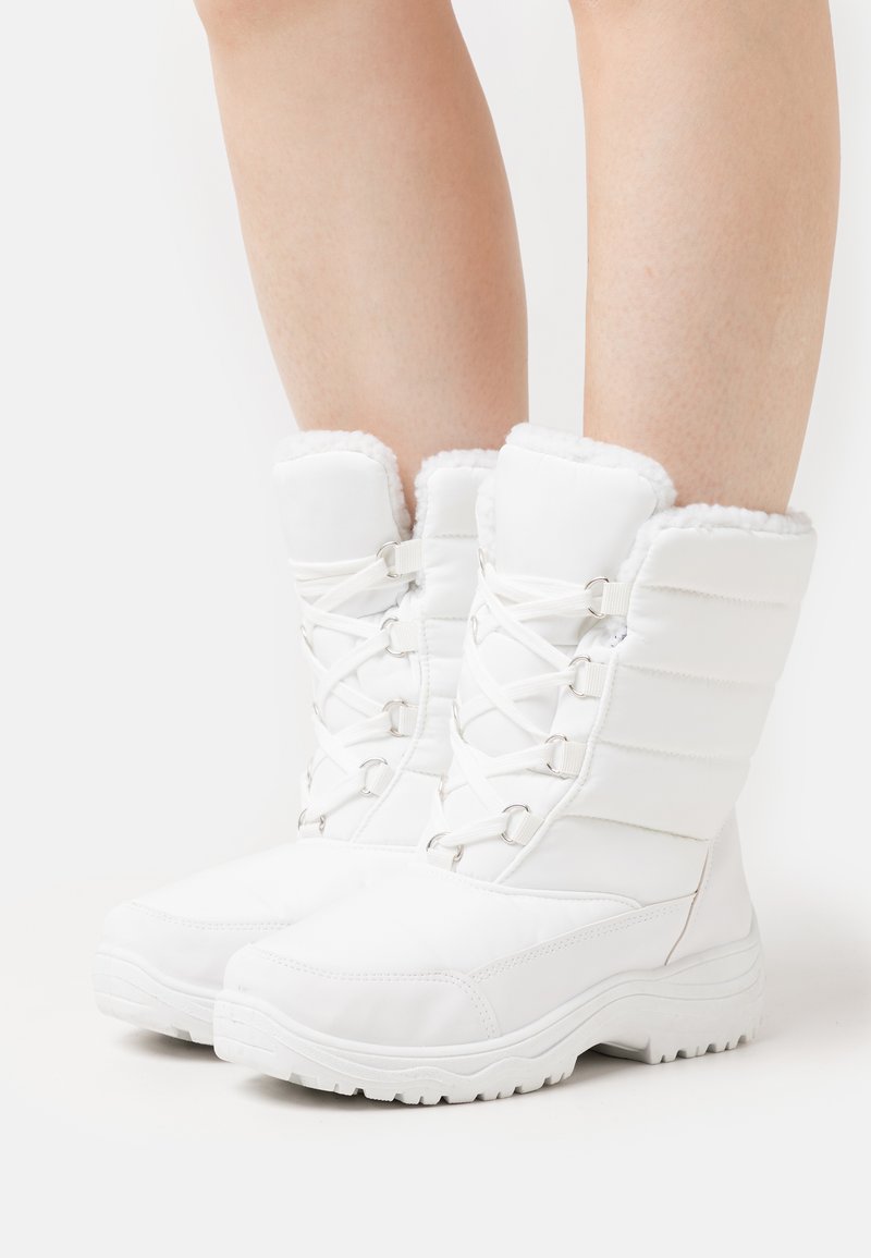 Des bottes d'hiver blanches présentent un design matelassé, une bordure en fausse fourrure et des semelles en caoutchouc flexibles. Devant à lacets renforcé avec des accents métalliques.
