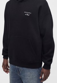 Μαύρο φούτερ Abercrombie & Fitch με μπροστινή τσέπη, φορεμένο από άτομο με μπλε τζιν, απεικονίζεται από τη μέση έως τα χέρια.