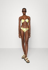 Biquíni floral amarelo com top bandeau detalhado com um anel central, combinado com parte de baixo de cintura baixa a condizer; modelo usa óculos de sol pretos e sandálias.