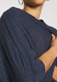 Frau trägt einen strukturierten dunkelblauen Pullover mit langen Ärmeln, hält den Stoff nahe ihrer Brust, Teils Gesicht und Arm sichtbar.