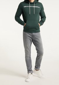 Felpa verde con tasca frontale, testo bianco e lacci; abbinata a jeans grigi e scarpe da ginnastica bianche. Tessuto morbido e vestibilità slim.