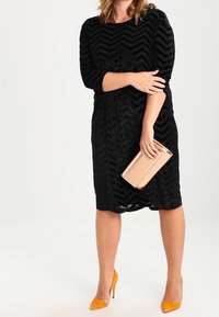 Black chevron-patterned dress with three-quarter sleeves, paired with yellow high heels and a rose gold clutch. Smooth texture, knee-length.