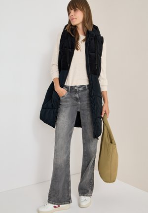Sort puffervest med en corduroy tekstur, lys beige sweater, grå wide-leg jeans og hvide sneakers, mens der holdes en olivengrøn taske.