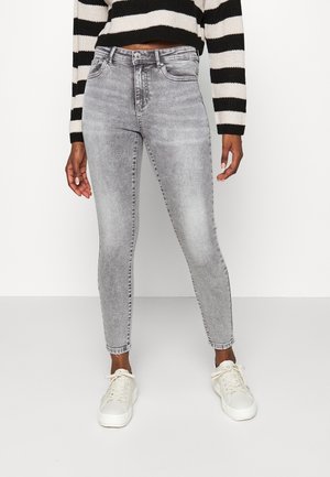Person, die hoch taillierte, ausgewaschene graue Skinny-Jeans, weiße Sneakers und einen schwarz-weiß gestreiften, bauchfreien Pullover trägt, vor einem einfarbigen Hintergrund steht.