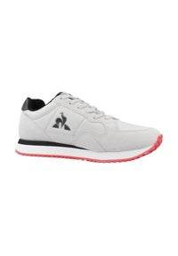 Zapatilla deportiva gris claro con acentos negros, material de ante, diseño con cordones y suela de goma rosa. Presenta un logo en el panel lateral.