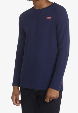 Osoba nosząca granatową długorękawową koszulkę Levi's z dzianiny waflowej z widocznymi czerwonymi naszywkami z logo Levi's na piersi i bocznym brzegu.